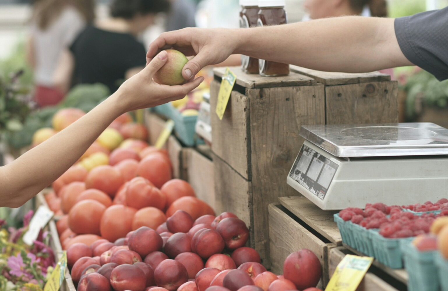 Marktszene: Eine offene Hand reicht einem Kunden einen Apfel – Sinnbild für den direkten Moment der Kundengewinnung am Point of Sale.